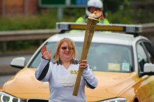 Claire waving and carrying the Olympic Torch followed by a car and police motorcyclist