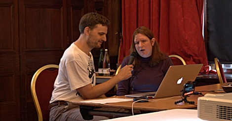 Man and woman speaking into a microphone, seated in front of a laptop and a projector