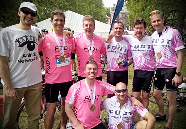 8 men arm in arm. 7 wearing pink cycling kit and one in a Aniridia Network UK tshirt
