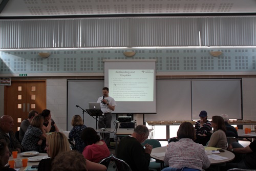 Man wearing ANUK t-shirt at lecturn talking to audience in a hall