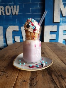 a pink milkshake in a jam jar style glass, with a straw and muffin sticking out the top and topped with cream and light blue and red sprinkles