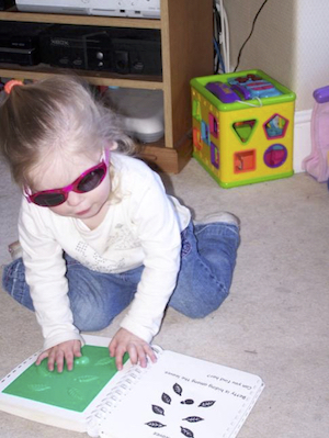 Small girl wearing dark glasses feeling a tactile book while knelling on the carpet