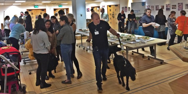 Delegate talking and eating lunch around exhibition stalls while man with guide dog passes by