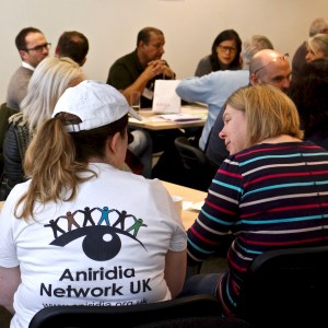 Two woman sitting with backs to the camera. One is wearing a Aniridia Network t-shirt. Other conference delegates are in the background.