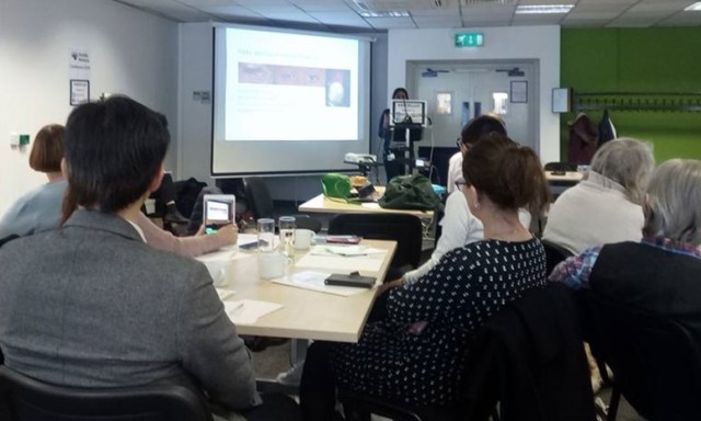 Seated delegates at tables, watching a presenter and big screen
