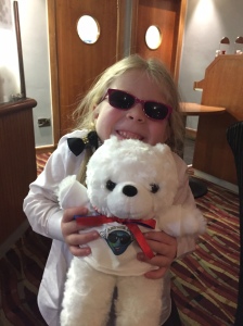 A young girl smiling and wearing sunglasses, while holding a white teddy bear. The bear has a red bow tied around its neck, and is wearing a white t-shirt featuring the Aniridia Day logo.