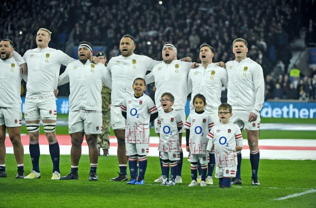 7 England men rugby players singing the national anthem on a grass pitch with 4 children in front of them