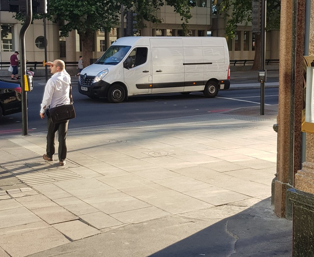 Man shielding his eyes from the bright sun at a pedestrian crossing that a van is driving across