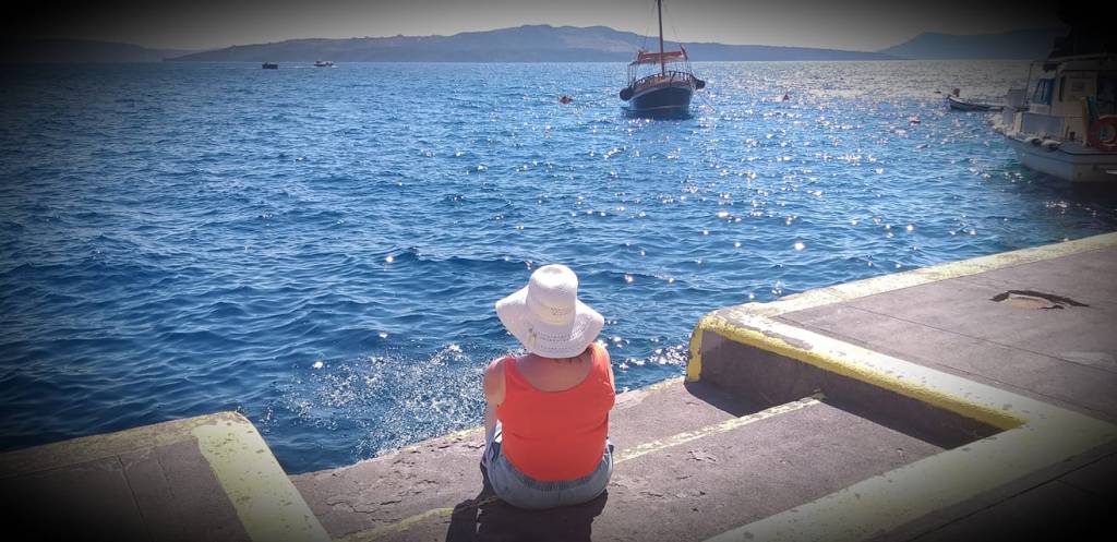 Eloise with their back to the camera, wearing a sun hat, sitting on a quayside, looking out at a very bright glitteirng sea.