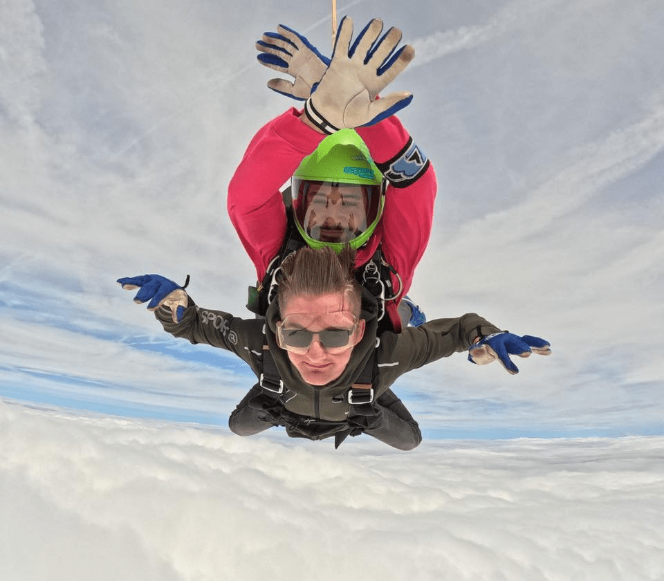 Oliver in a black jump suit strapped under an instructor falling through mid air above the clouds arms outstretched, looking at the camera