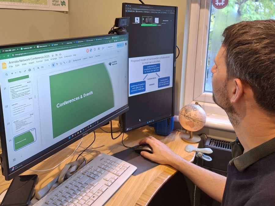 James running an Aniridia Network online conference from his home office. He is using 2 monitors. The left is editing the main slide deck showing 'Conferences and Events' and the right has an in progress talk on Zoom