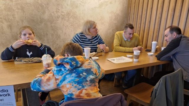 2 children, 1 woman, 2 men eating and talking at a coffee shop table that has a Aniridia Network poster and white cane on it.